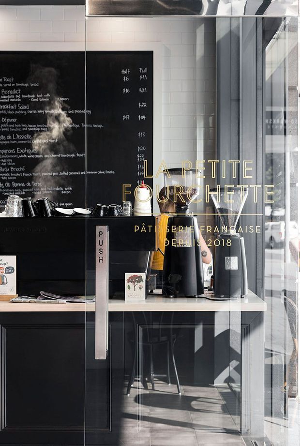 A contemporary French-inspired patisserie or café interior viewed through a glass entrance. The compact service counter features black cabinetry, a pale timber benchtop, coffee equipment, cups, and small display items. Behind it is a black handwritten chalkboard menu framed by white subway tile walls. Strong sunlight enters through the glazed storefront, creating reflections and shadows, while gold lettering on the glass identifies the venue.