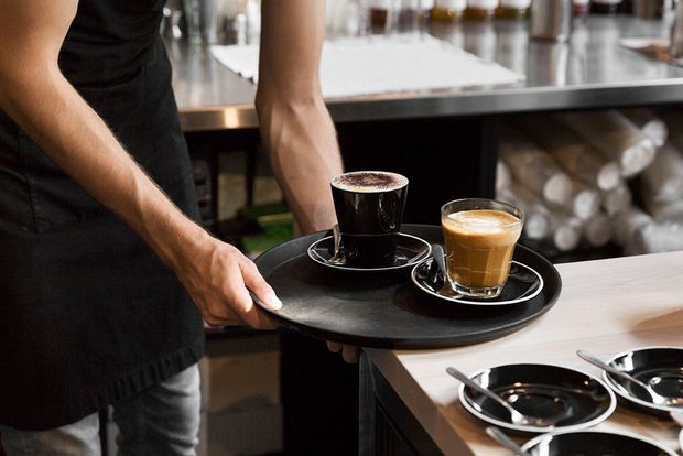 Close-up of a café or commercial hospitality workspace where a server in a black apron carries a black serving tray with two coffees. Stainless-steel work surfaces, shelving, and stacked crockery are visible in the background, with additional cups and saucers on a light timber-toned counter.
