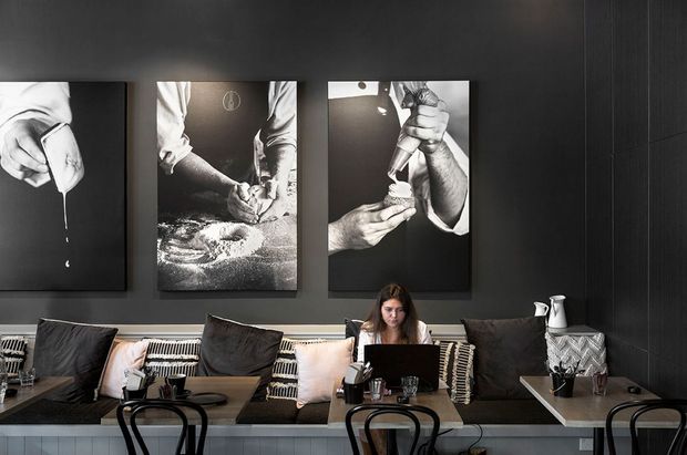 A moody, modern hospitality interior with a long built-in banquette upholstered in dark and patterned textiles, small dining tables, and black bentwood-style chairs. A person works at a laptop, while large monochrome culinary photographs decorate the dark textured wall behind the seating.