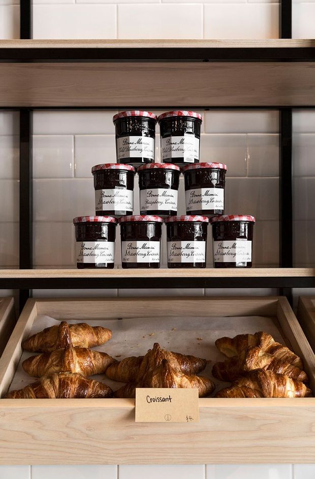 Close-up of a bakery or café display with wooden shelving and trays holding croissants, alongside stacked jars of preserves. White subway-tile backsplash, dark metal shelf framing, and warm timber joinery create a simple, rustic commercial fit-out.