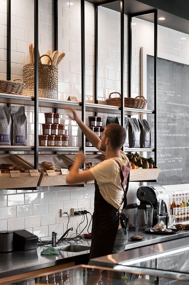 A compact, modern café or bakery serving area with open black metal shelving, woven baskets, packaged goods, bread displays, a stainless-steel work counter, sink, coffee equipment, and a large chalkboard menu. White subway-tile walls and dark framing create an industrial-inspired commercial fit-out.