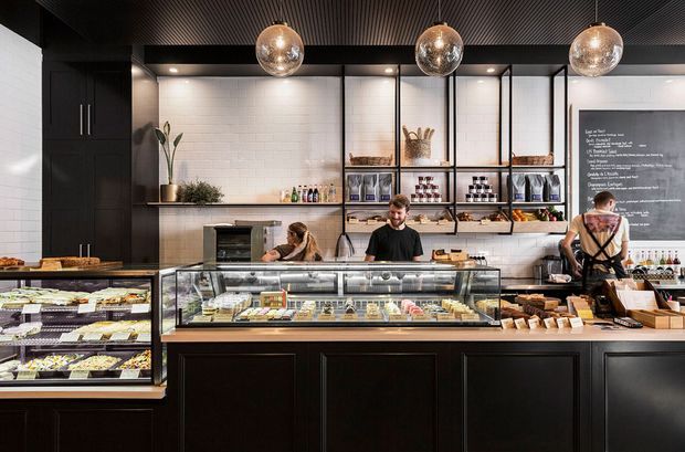 A contemporary retail food counter or café interior with dark timber cabinetry, glass display cases filled with prepared foods, open black-framed shelving, white brick wall tiles, and a dark slatted ceiling. Three spherical pendant lights illuminate the service counter, with staff working behind it and a chalkboard menu at right.