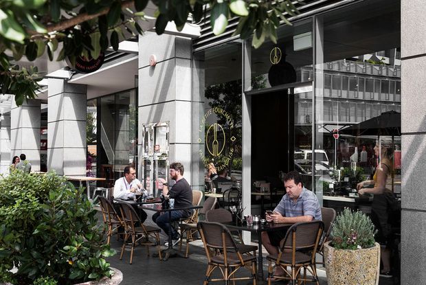 Street-level view of a contemporary cafe or restaurant with outdoor dining on a paved terrace. Large glazed shopfronts, dark metal framing, light grey stone or concrete-clad structural columns, and black awnings define the facade. Customers sit at small tables with woven chairs, while shrubs and potted plants soften the frontage.