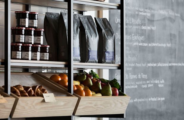 Close-up of a contemporary retail or café display with pale plywood produce bins and open shelving. The shelves hold jars, packaged goods, bread, and fresh fruit, while a large dark chalkboard-style wall with handwritten menu or product text forms the background. Black metal framing and a muted industrial material palette create a modern shop interior.