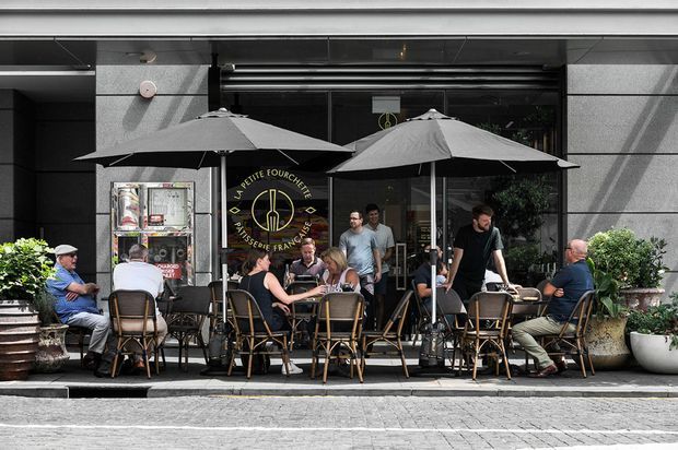 Street-level view of a French-style café or patisserie frontage with outdoor dining tables, wicker café chairs, large dark umbrellas, potted plants, and customers seated along a paved sidewalk. The façade has dark glazing, grey stone or concrete surfaces, and visible French café signage.