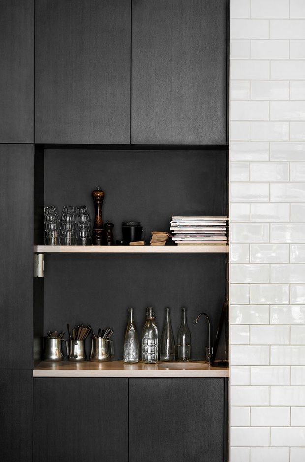 Close-up of a dark, built-in kitchen or beverage cabinetry niche with matte charcoal cabinetry, open timber shelves, glassware, bottles, utensils and a small sink tap. A glossy white subway-tile wall forms the right side of the composition.