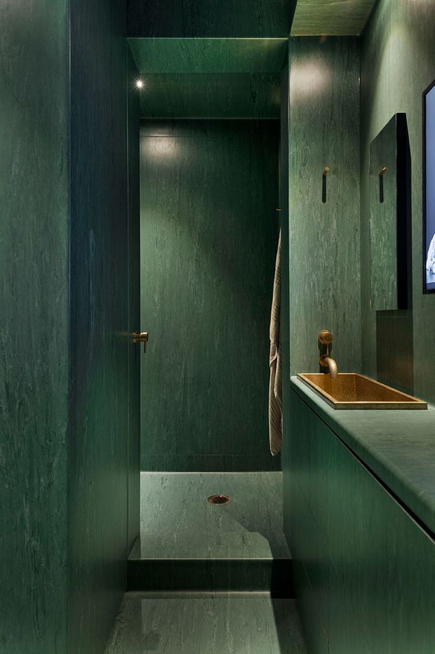 A narrow, moody bathroom clad almost entirely in deep green textured surfaces, including the walls, ceiling, floor, and built-in vanity. The compact wet-room area has a step-up shower floor with a central drain, a towel hanging beside the shower, and a long vanity with a bronze-toned rectangular basin and matching brass/bronze tapware. A mirror and partially visible wall-mounted screen appear on the right. The space has dramatic low-level lighting and a highly tonal, immersive finish.
