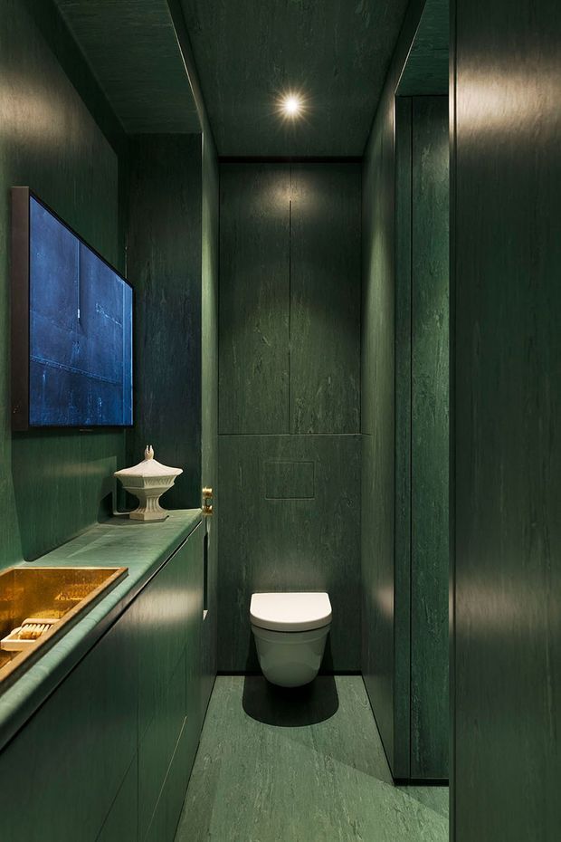 A compact, dramatic powder room clad almost entirely in deep forest-green textured finishes. The narrow room contains a wall-mounted toilet, a floating vanity with a warm brass/gold basin, a small ornate white vessel, dark green stone-like flooring, integrated cabinetry, a wall-mounted screen or artwork, and a single recessed ceiling light.