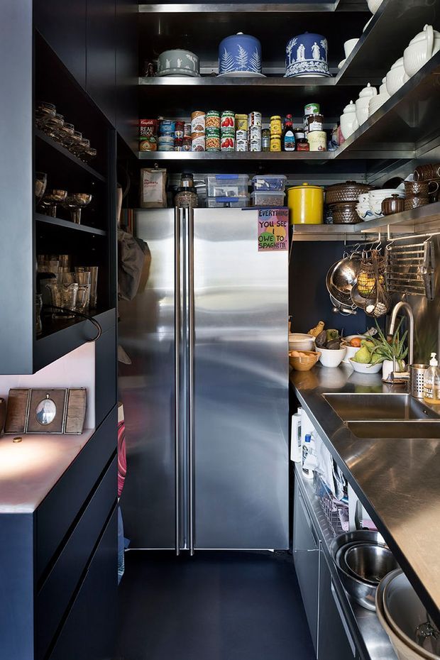 A compact, highly organized galley kitchen with dark built-in cabinetry, open shelving, stainless-steel appliances and countertops, a double-door refrigerator, sink, hanging cookware, and extensive pantry storage. Decorative ceramics and food containers are displayed on the shelves, with a small handwritten note on the refrigerator.