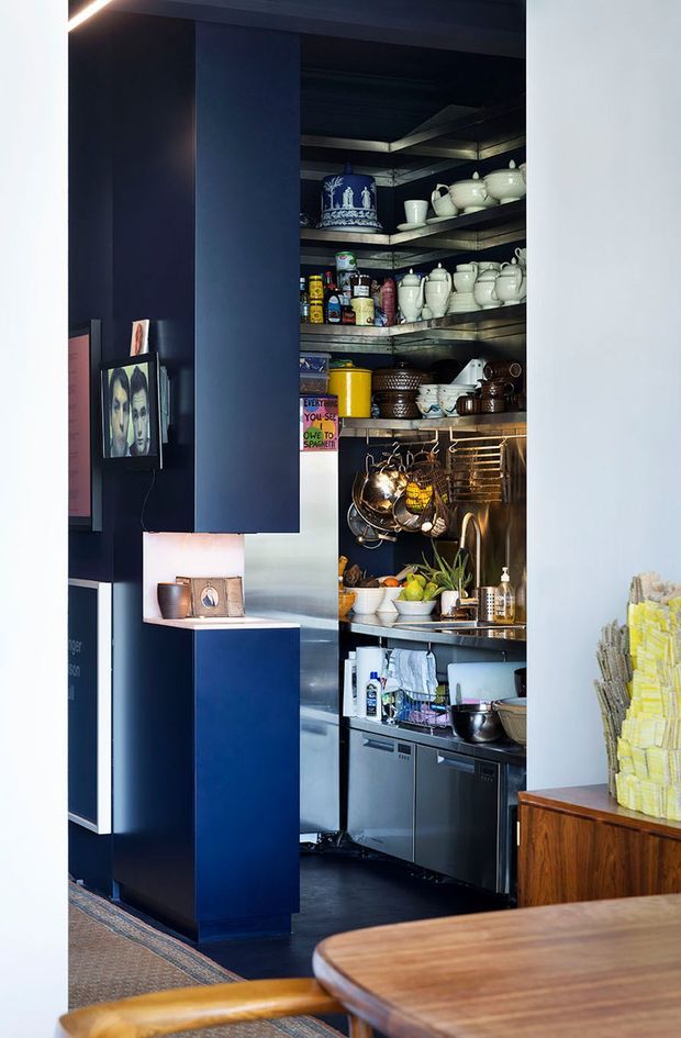 A compact, dark-toned kitchen or pantry alcove with open industrial-style shelving densely stocked with crockery, cookware, jars and pantry supplies. Deep navy built-in cabinetry and partitions frame the space, with stainless-steel appliances and a sink below. A timber dining table, framed artwork and warm task lighting are visible in the foreground and adjacent area.