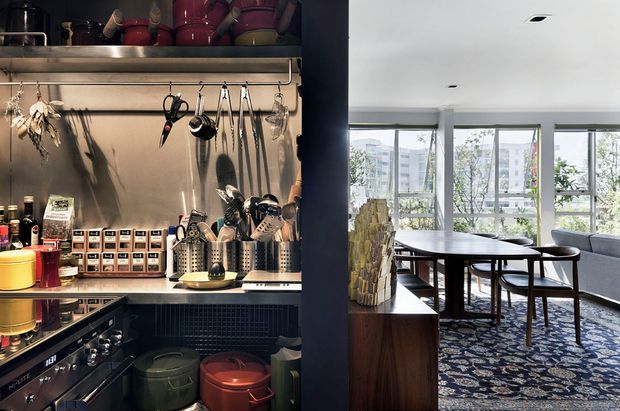 A split view of a compact, highly organized kitchen and dining area. The left side shows open stainless-steel shelving filled with cookware, utensils, jars, and countertop appliances, with dark cabinetry and colourful cookware below. The right side shows a bright dining nook with a dark timber table and chairs, patterned rug, stone-clad feature column, and large corner windows overlooking an urban setting.