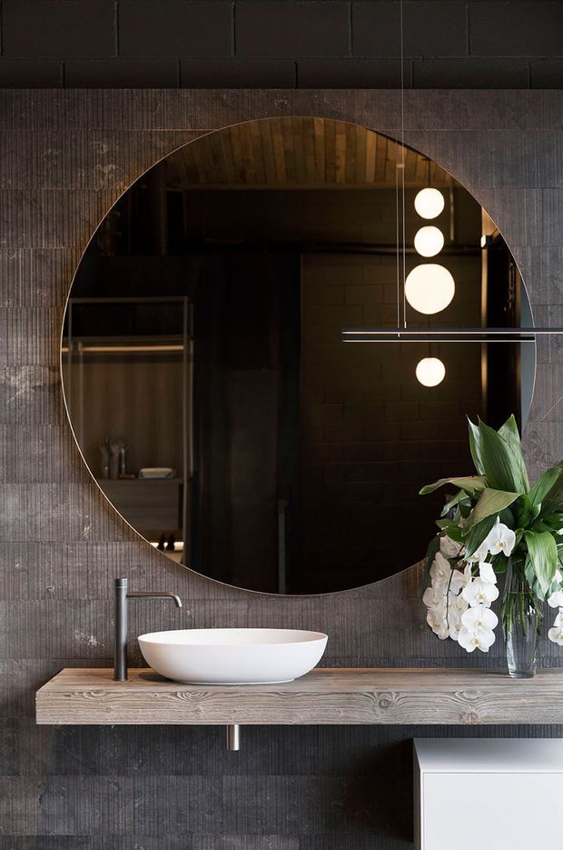 Moody contemporary bathroom vanity with a large round mirror, white vessel basin, dark vertically textured wall finish, timber floating vanity shelf, matte black tapware, and spherical pendant lights reflected in the mirror. A floral arrangement adds a natural accent, while white cabinetry is partially visible below.