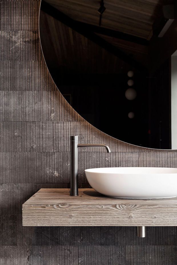 A moody, contemporary bathroom vanity featuring a white countertop basin, brushed metal wall-mounted or deck-mounted tap, and a thick floating timber vanity shelf. The walls are clad in dark vertically textured tiles, while a large rounded mirror reflects a dark timber ceiling and pendant lights.