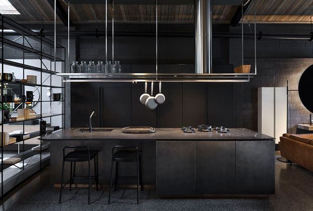 Dark, sophisticated industrial-style kitchen dominated by a large island with a dark stone benchtop and dark built-in cabinetry. Black metal open shelving, suspended metal framing, hanging cookware, exposed timber ceiling boards, stainless-steel ventilation ducting, and a textured dark wall create a loft-like aesthetic. A leather sofa and adjacent living area are visible at the right.