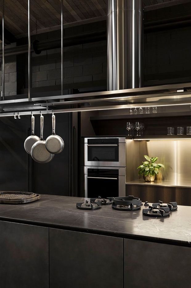 A dramatic, dark-toned industrial kitchen with a large stone island and integrated gas cooktop, black cabinetry, stainless-steel double ovens, metallic rangehood ducting, and open shelving displaying glassware. Hanging pans, exposed dark blockwork, visible ceiling structure, and warm under-shelf lighting reinforce the utilitarian aesthetic.