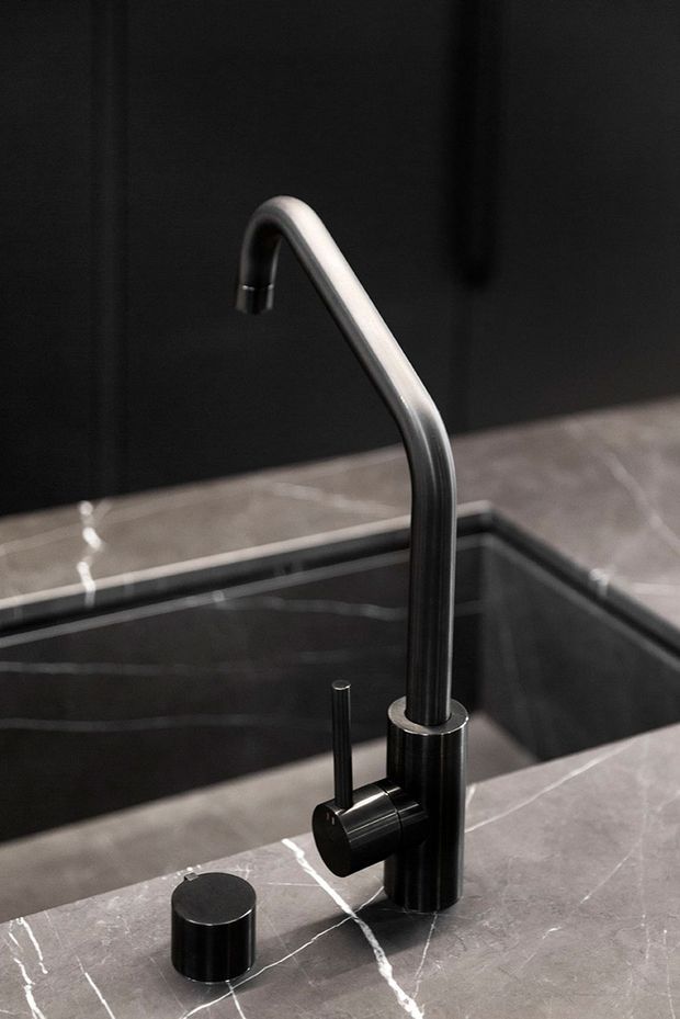 Close-up product-style image of a sleek black and brushed-metal kitchen tap mounted beside a dark undermount sink. The tap and separate cylindrical control sit on a grey marble-look benchtop, with dark cabinetry or wall surfaces behind. The composition is minimalist, high-contrast, and refined.