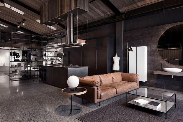 An open-plan loft-style interior combining a living room and kitchen. The space features dark industrial walls, exposed black services and ductwork, a timber-lined ceiling, polished concrete or terrazzo flooring, a dark kitchen island with a large stainless-steel extraction hood, and a brown leather sofa. A glass coffee table, sculptural lighting, metal shelving, and a partially visible bathroom vanity add refined contemporary furnishings. The room has a dramatic, moody atmosphere with strong industrial character.