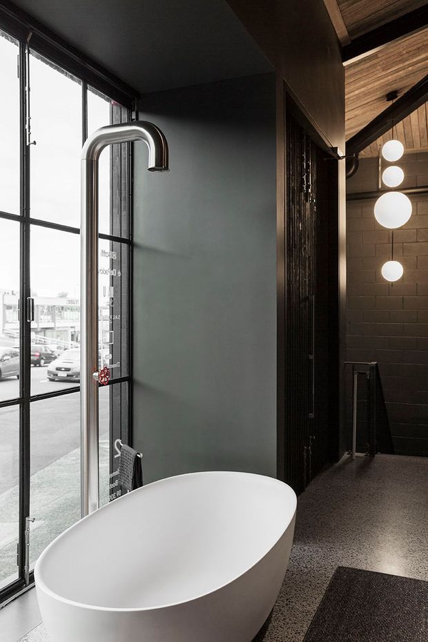 A dramatic, contemporary bathroom featuring a freestanding white oval bathtub beside full-height black-framed glazing. A tall curved chrome bath filler rises from the floor, while a dark partition opens toward an adjacent space with charcoal brick walls, exposed timber ceiling elements, and spherical pendant lights. The room has dark grey walls and polished speckled flooring, creating a moody industrial atmosphere.