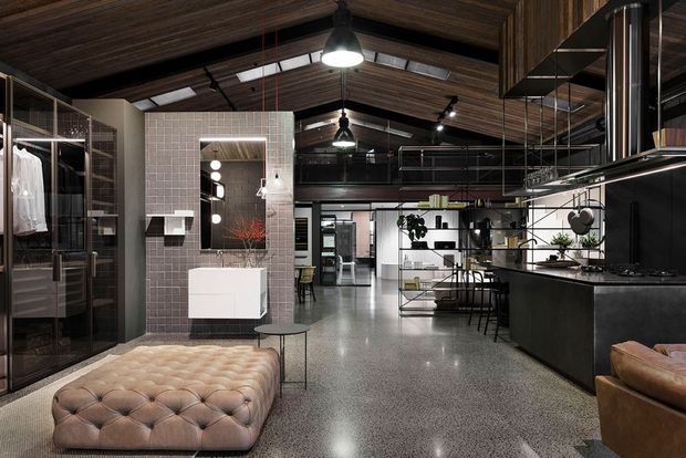 A spacious industrial-style open-plan interior combining a kitchen, living area and walk-in wardrobe. The room features dark timber-lined pitched ceilings with exposed beams, polished concrete or terrazzo-like flooring, black metal-framed glazing and shelving, a dark kitchen island, and a tufted leather ottoman in the foreground. A tiled bathroom zone with a wall-mounted basin and mirror is visible at the rear left.
