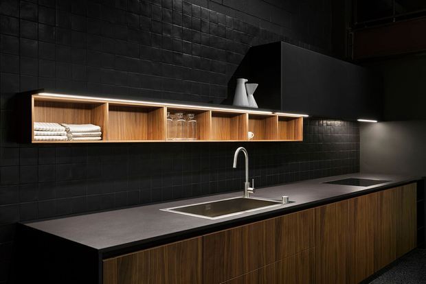 A dark, sophisticated kitchen or kitchenette featuring matte black tiled walls, a long charcoal benchtop, warm timber cabinetry, and an open timber display shelf with folded towels and glassware. A stainless-steel sink and minimalist gooseneck tap sit beneath understated linear lighting, creating a moody, refined atmosphere.