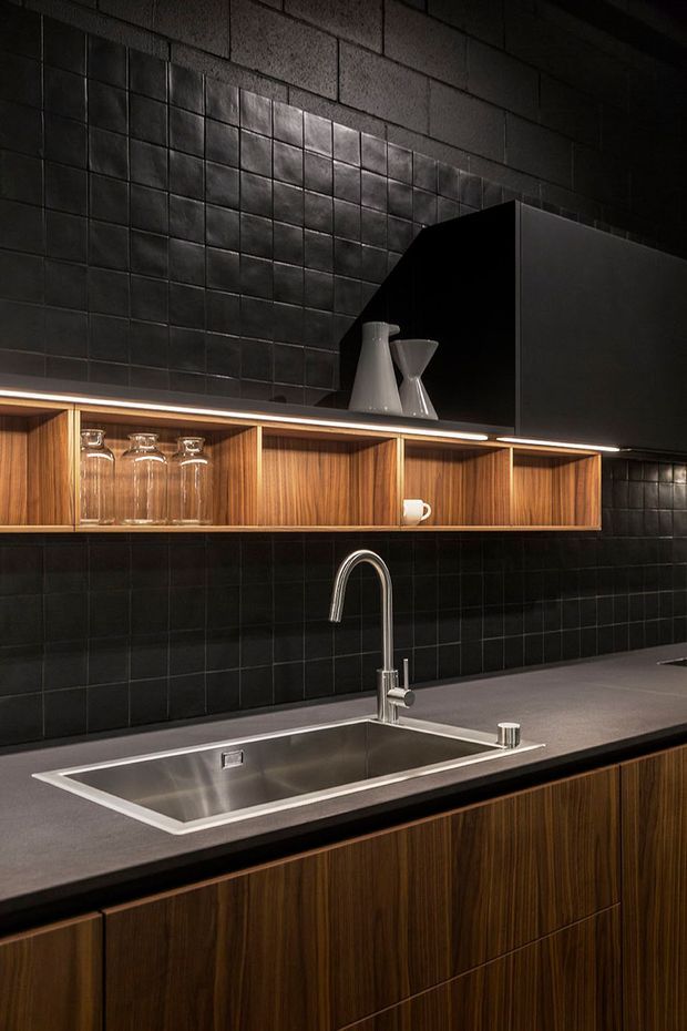 A dark, sophisticated contemporary kitchen featuring black square tiled walls, warm timber open shelving and cabinetry, a long grey stone benchtop, and an integrated stainless-steel sink with a curved chrome tap. Under-shelf lighting highlights glassware and ceramic vessels, while a large dark rangehood or overhead cabinet occupies the upper right.