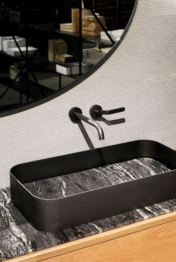 Close-up of a refined bathroom vanity featuring a matte black vessel basin with rounded corners, wall-mounted black tapware, and a dramatic black-and-white natural stone benchtop. The wall is clad in a fine light textured tile, with a large curved mirror reflecting dark shelving and stacked objects. A warm timber vanity front provides contrast.