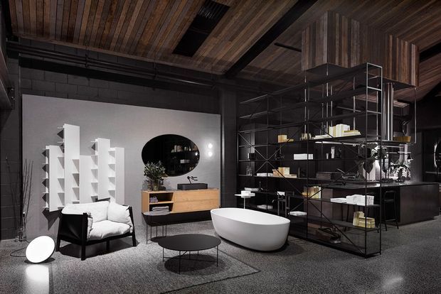Dark, dramatic open-plan interior showroom or living space with a warm timber-lined ceiling, exposed black structural and service elements, grey walls and carpeted flooring. The room combines a white freestanding bathtub, black metal open shelving, a timber sideboard, sculptural white display shelving, lounge seating, and low coffee tables, creating an industrial bathroom-and-living display.