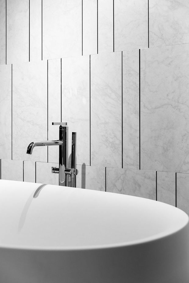 Close-up bathroom detail featuring a freestanding white bathtub in the foreground, a polished chrome bath mixer and hand shower, and a wall clad in large vertically arranged pale marble-effect tiles with dark grout lines. The monochromatic, high-contrast composition highlights the sculptural tub and contemporary fixtures.