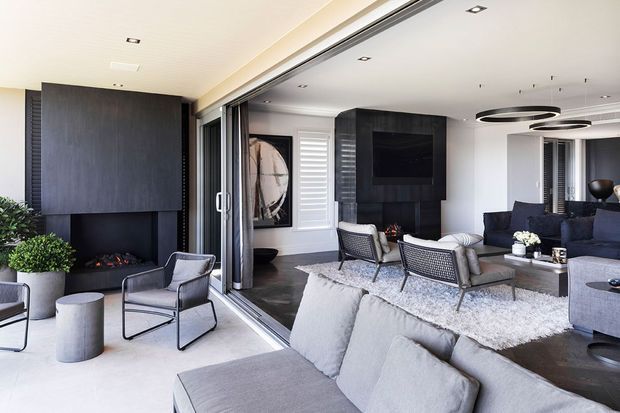 Contemporary open-plan living room opening onto an outdoor terrace through wide sliding glass doors. The space features a large grey sectional sofa, dark stone or timber feature walls, a built-in fireplace, wall-mounted television, modern pendant lighting, and neutral furnishings. Outdoor lounge chairs and potted greenery are visible on the terrace.