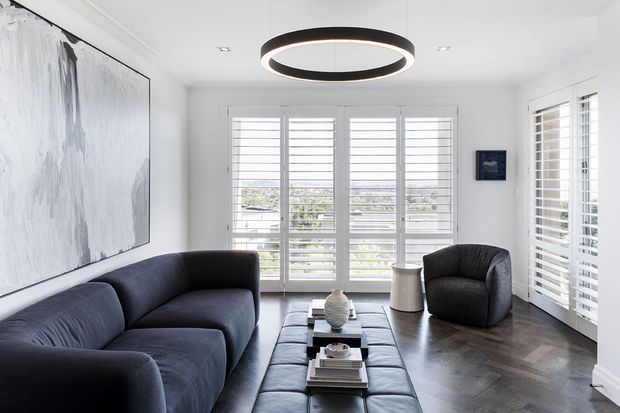 Bright, refined living room with dark upholstered sofas and ottoman seating, white walls, extensive plantation shutters, dark herringbone timber flooring, and a contemporary circular pendant light. Large windows provide an airy outlook, while abstract wall art and restrained styling create a sophisticated monochromatic interior.