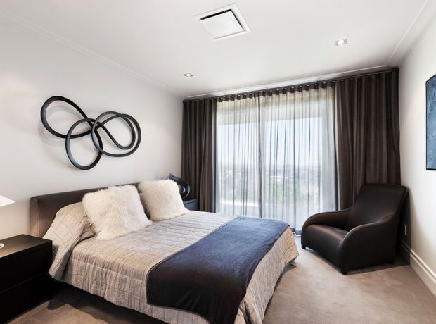 A contemporary bedroom with a neatly made bed, layered neutral and dark bedding, a black upholstered armchair, dark full-height curtains, and sheer curtains covering a large window. The room has white walls and ceiling, beige carpet, recessed lighting, and abstract circular wall decor.