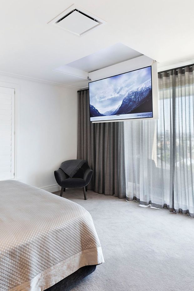 Bright, refined bedroom with a large upholstered bed, pale carpet, white walls and ceiling, sheer and dark layered curtains, and a black lounge chair. A wall-mounted television is integrated into a ceiling-mounted retractable lift or concealed cabinetry feature. A large window provides filtered daylight, while a ceiling air-conditioning vent is visible above.