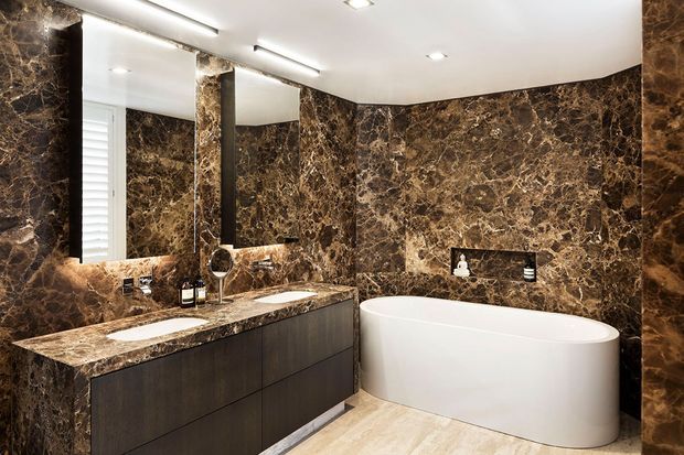 Luxurious contemporary bathroom featuring extensive dark brown veined stone wall cladding and matching vanity benchtop, a double-sink floating dark timber vanity, large mirror, recessed shelf, and a freestanding white oval bathtub. Recessed ceiling lighting creates a refined, high-contrast atmosphere.