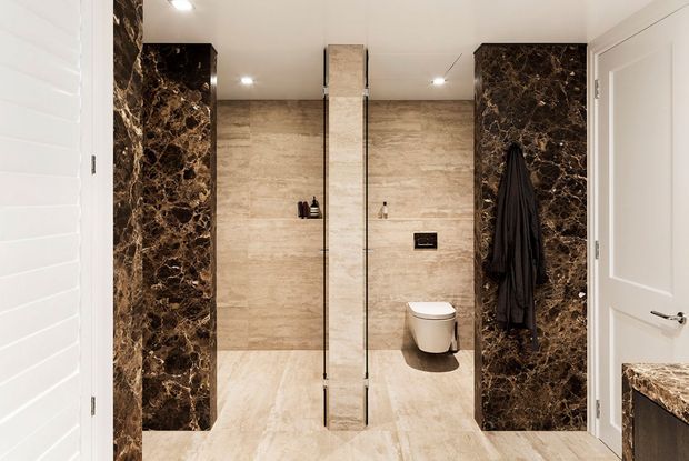 A refined contemporary bathroom with a walk-in shower enclosed by clear glass panels, a wall-hung toilet, and full-height beige stone-look wall and floor tiling. Dramatic dark brown marble-look feature panels frame the shower and toilet areas, with white doors and recessed ceiling lighting.