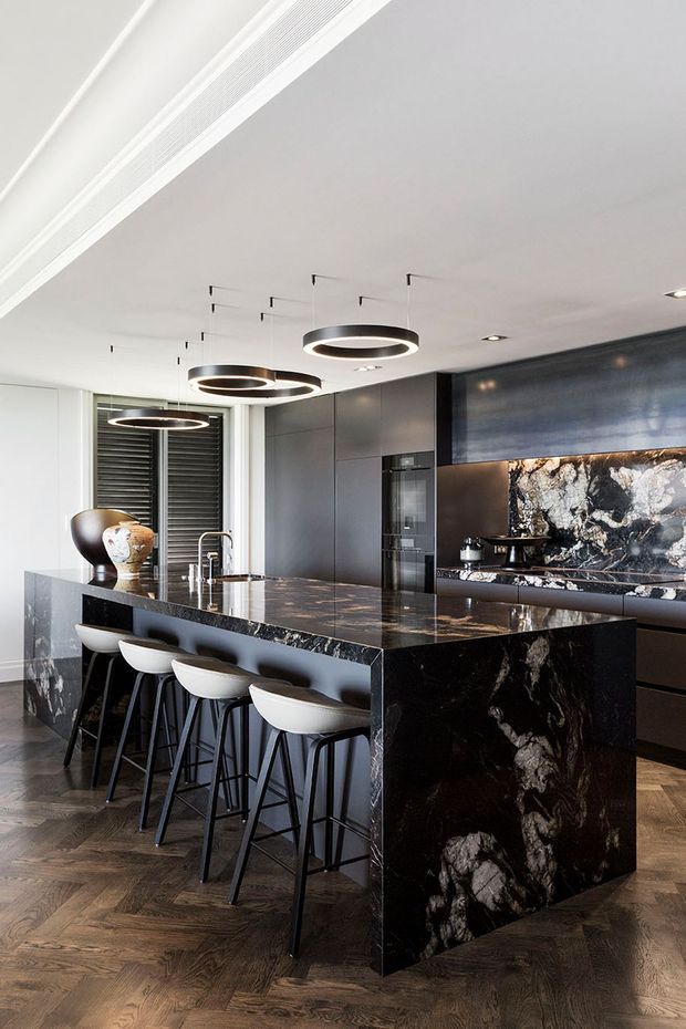 A sophisticated contemporary kitchen centered on a large dark marble island with waterfall ends and seating for four. Matte dark cabinetry, integrated appliances, a dramatic marble splashback, white walls, herringbone timber flooring, and sculptural black-and-white pendant lights create a high-contrast, luxurious interior.