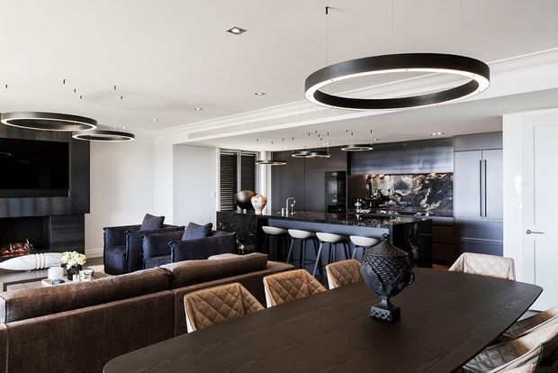 A sophisticated open-plan living, dining and kitchen interior. The room features dark built-in cabinetry, a black stone kitchen island, sculptural circular pendant lights, a large dark dining table with upholstered chairs, a sectional sofa, and a fireplace with television wall. The palette is predominantly charcoal, black, grey and warm brown, creating a refined contemporary atmosphere.