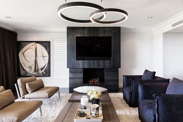 A refined contemporary living room featuring a dark timber or charcoal feature wall with a built-in television and fireplace, plush navy sofas, tan leather lounge chairs, a large textured rug, and a long timber coffee table. Sculptural circular pendant lights create a striking focal point, while artwork, plantation shutters, and full-height dark curtains complete the space.