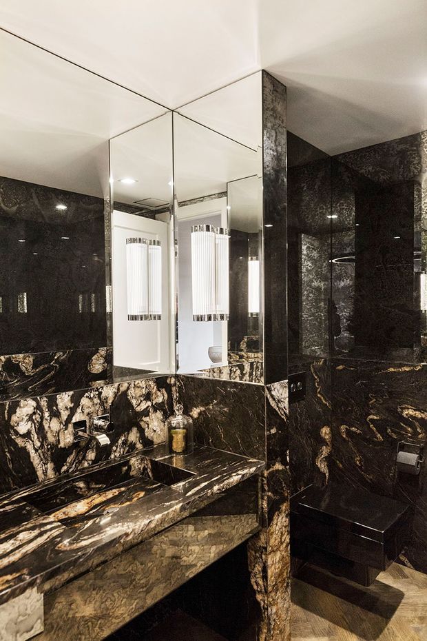 A dramatic, luxurious bathroom clad almost entirely in richly veined dark stone. The image shows a stone vanity with integrated basin, wall-mounted tapware, large mirrored panels, sculptural vertical wall lights, and a partially visible enclosed shower area. The dark marble-like surfaces create a high-contrast, opulent atmosphere.