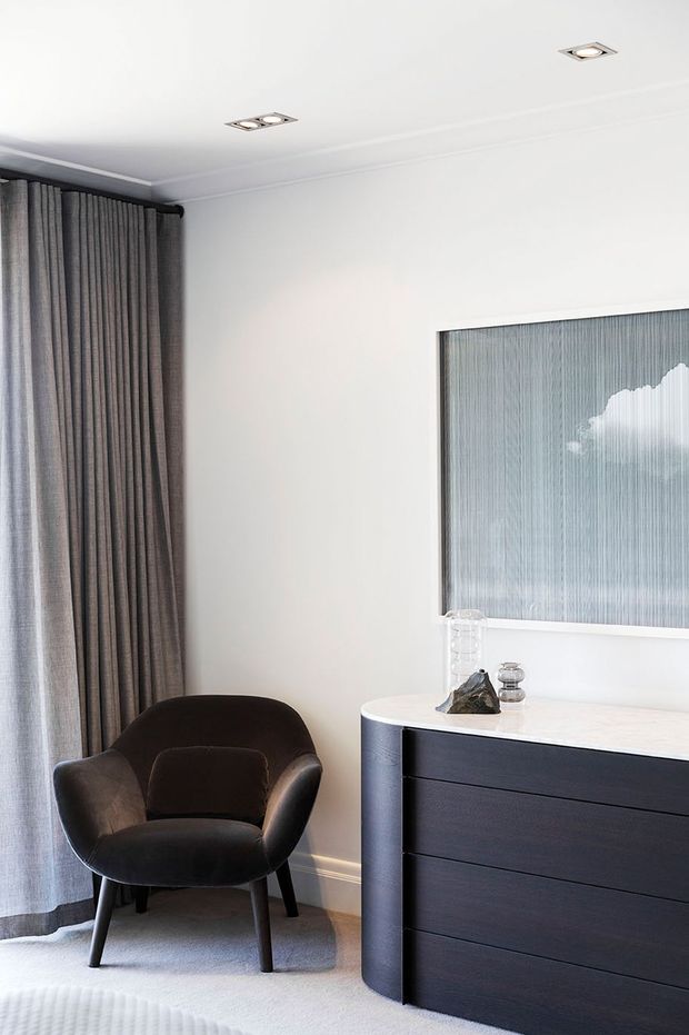 A refined bedroom corner featuring a dark upholstered lounge chair beside a dark built-in chest of drawers with a pale stone or solid-surface top. Full-height grey curtains frame the window, while a large framed artwork and recessed ceiling spotlights complete the calm, monochromatic interior.