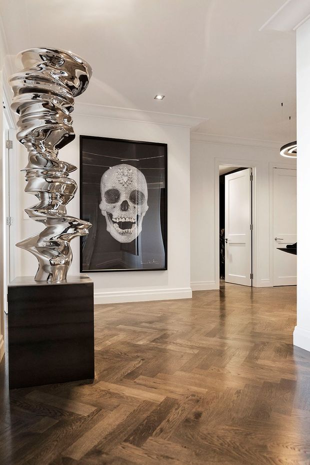 Elegant residential entry hall or gallery with a polished herringbone timber floor, white paneled walls and ceiling trim, and multiple white interior doors. A dramatic reflective silver sculptural artwork stands on a black plinth beside a large framed skull artwork. The space is bright, refined, and art-focused, with contemporary lighting and classic architectural detailing.