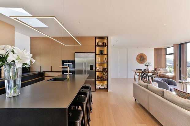 Open-plan contemporary kitchen and living area with a large dark island and integrated seating, warm timber cabinetry, stainless-steel appliances, open shelving, pale timber flooring, and linear pendant lighting. A dining and lounge area extends toward large windows with a distant urban view.