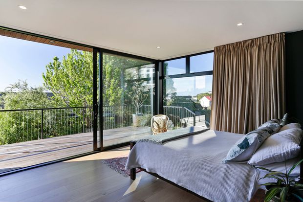 Bright contemporary bedroom with a large bed, soft neutral bedding, full-height corner glazing, and sliding doors opening to a timber deck and leafy outlook. Floor-to-ceiling curtains and dark window frames contrast with the light timber-toned floor and white ceiling.