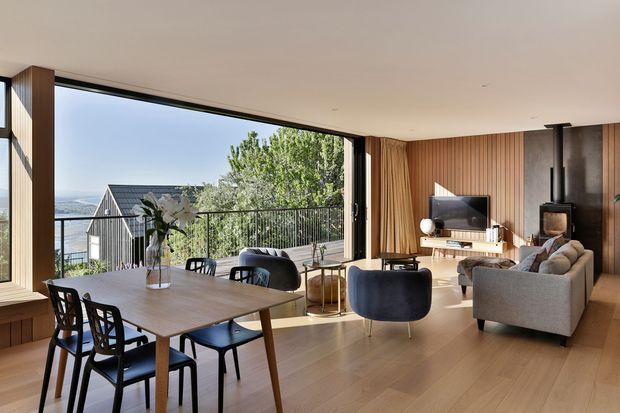 A bright, open-plan living and dining room with full-height glazing opening to a balcony or terrace. The space features warm timber flooring and vertical timber wall panelling, a large dining table with dark chairs, a curved dark lounge chair, a grey sofa, a television, and a compact wood-burning stove. Mountain and water views are visible beyond the glazing.