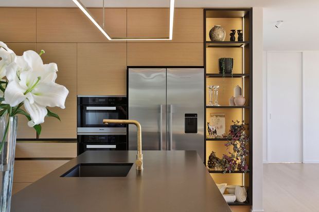 Contemporary kitchen featuring warm timber cabinetry, integrated black appliances, stainless-steel refrigerator, dark stone island benchtop with undermount sink and curved brass tapware, and a backlit open display shelving unit. A bright white hallway or adjoining living area is visible beyond.