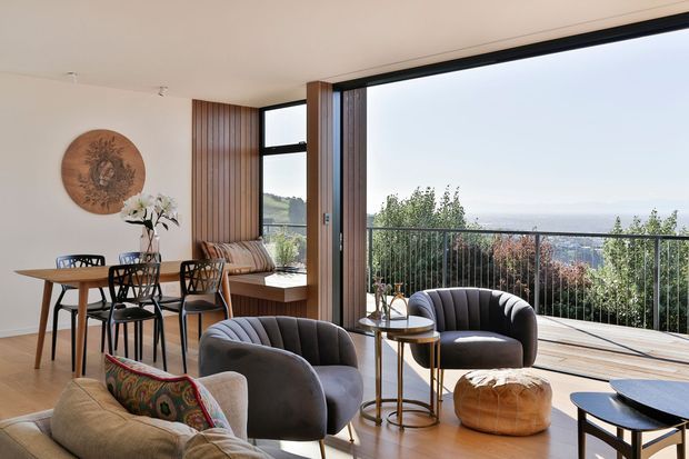 A bright, open-plan living and dining area with expansive floor-to-ceiling glazing opening onto a balcony or terrace with a metal balustrade and distant landscape views. The room features warm timber flooring, a white ceiling and walls, vertical timber wall panelling, a compact dining table with mixed chairs, curved upholstered lounge chairs, a sofa, side tables, and a built-in window seat. Natural light and warm neutral furnishings create a refined, inviting atmosphere.