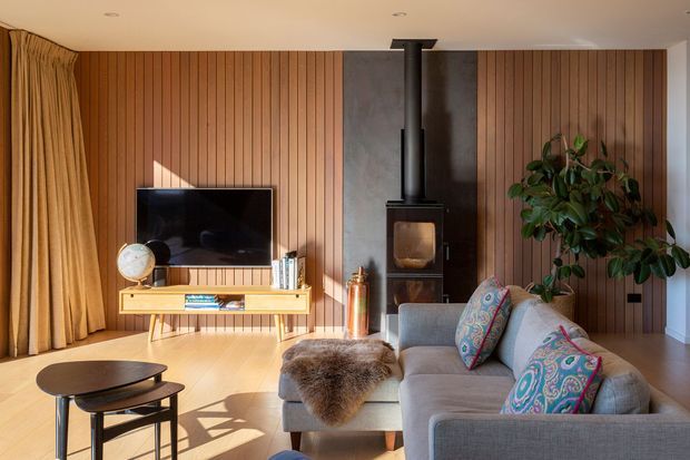 Warm, inviting living room with vertical timber wall panelling, a wall-mounted television on a slim timber media console, a freestanding black wood-burning stove with flue, grey sofa and ottoman, patterned cushions, round nesting coffee tables, and full-height beige curtains. Sunlight creates strong shadows across the timber floor, with a large indoor plant adding greenery.