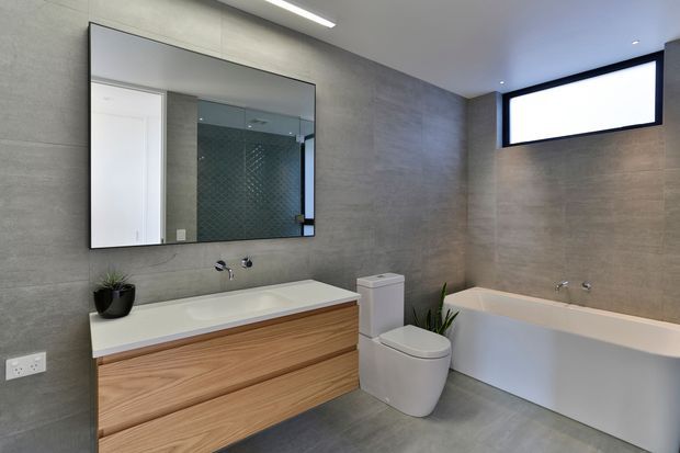 Contemporary bathroom featuring large-format grey wall and floor tiles, a floating natural timber vanity with integrated white basin, wall-mounted tapware, a frameless mirror, white toilet, and a white built-in bathtub. A small high window provides natural light, while a recessed linear ceiling light adds illumination.
