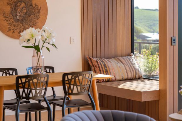 Warm, light-filled dining area beside a large glazed opening, featuring a timber dining table, black geometric-back chairs, a built-in timber window seat with striped cushions, vertical timber wall panelling, and a round carved wall artwork. A green landscape is visible outside.