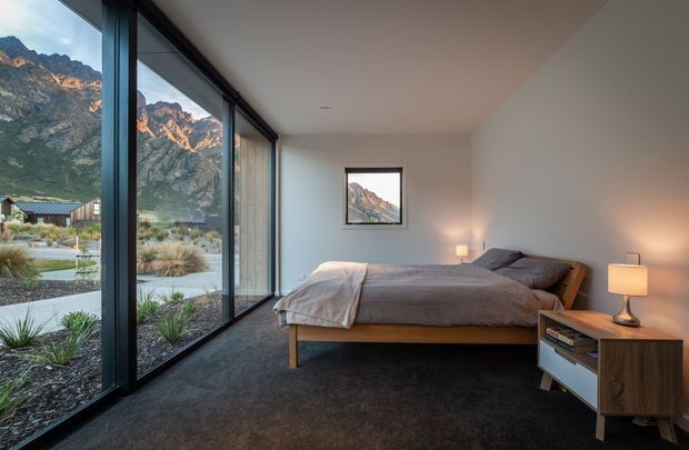 A minimalist bedroom with a low timber bed and grey bedding, dark carpet, white walls, and simple bedside lighting. Full-height black-framed glazing runs along one side, providing expansive views of rugged mountain terrain and a landscaped exterior. The room has a calm, restrained atmosphere and strong indoor-outdoor connection.