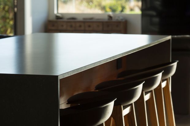 Close-up interior view of a large kitchen island or breakfast bar with a dark matte benchtop, timber-toned front panel, and several sculptural dark stools with timber legs. A sideboard and windows are softly visible in the background, with warm natural light creating a refined, contemporary atmosphere.